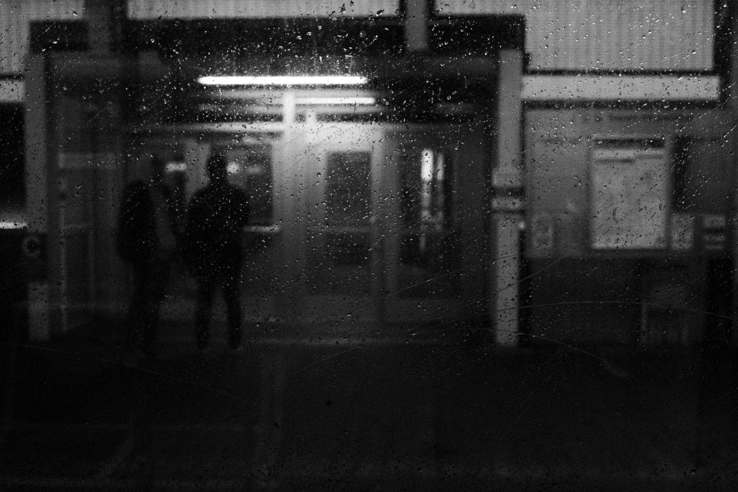 Rain-speckled window blurring the view of two silhouetted figures standing outside a train station entrance, illuminated by overhead lights. Rain-speckled window blurring the view of two silhouetted figures standing outside a train station entrance, illuminated by overhead lights.