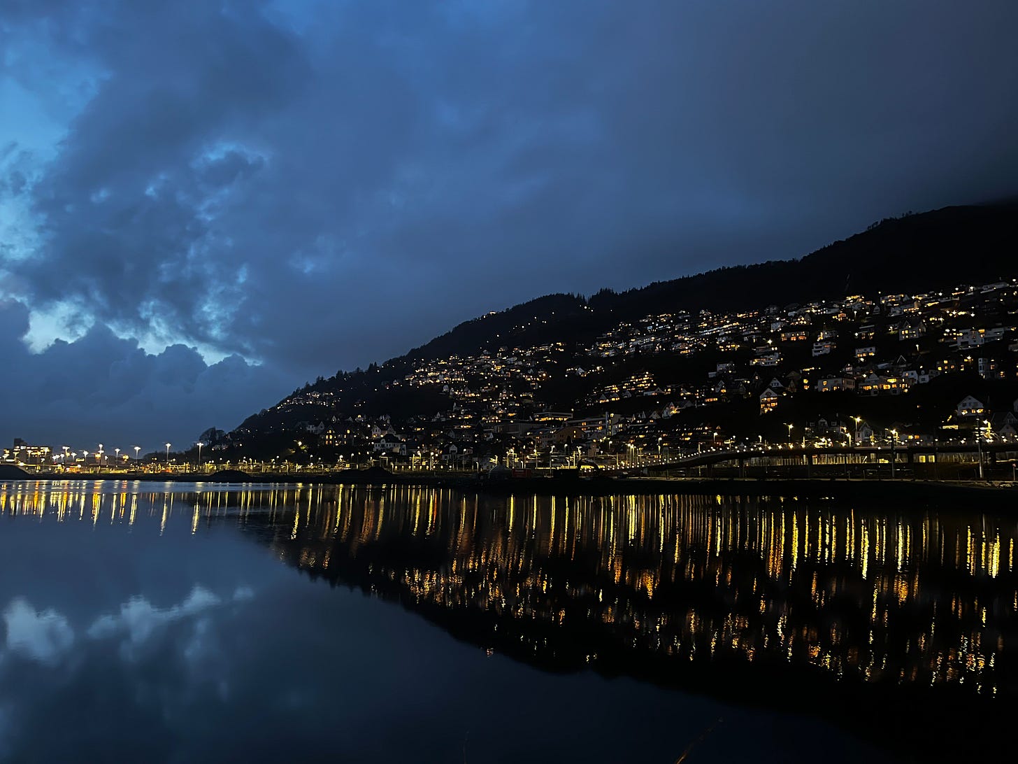 The silhouette of a hillside city overlooking a lake at night. The lights in the city glow softly and reflect in the lake