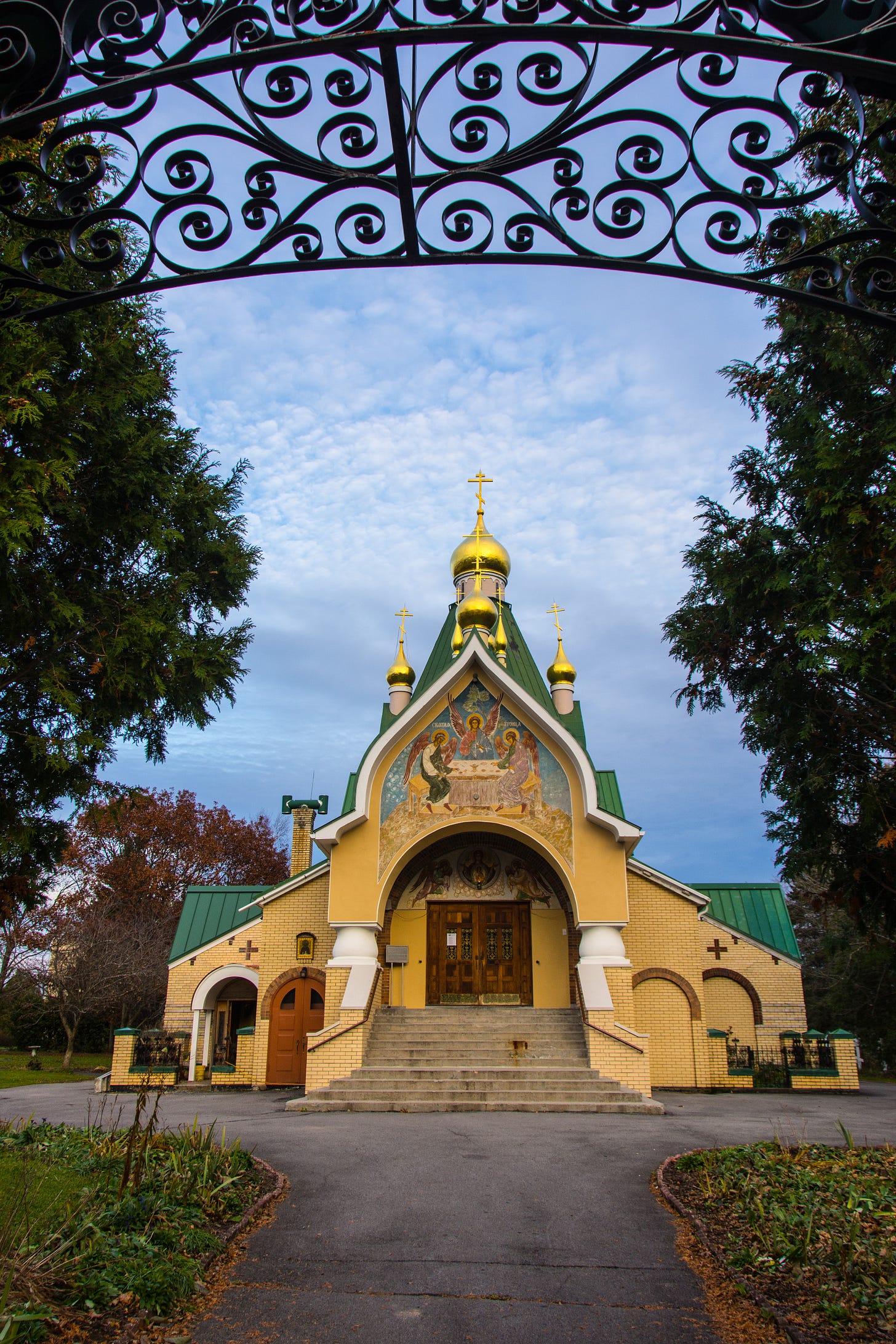 Front facade of a yellow-brick church with green roofs and gold onion domes topped with crosses, seen through a wrought-iron gate arch, with a mosaic above the entrance.