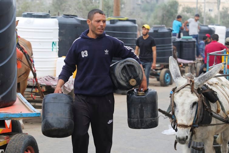 A man carrying three large plastic jugs walks next to a donkey cart A man carrying three large plastic jugs walks next to a donkey cart