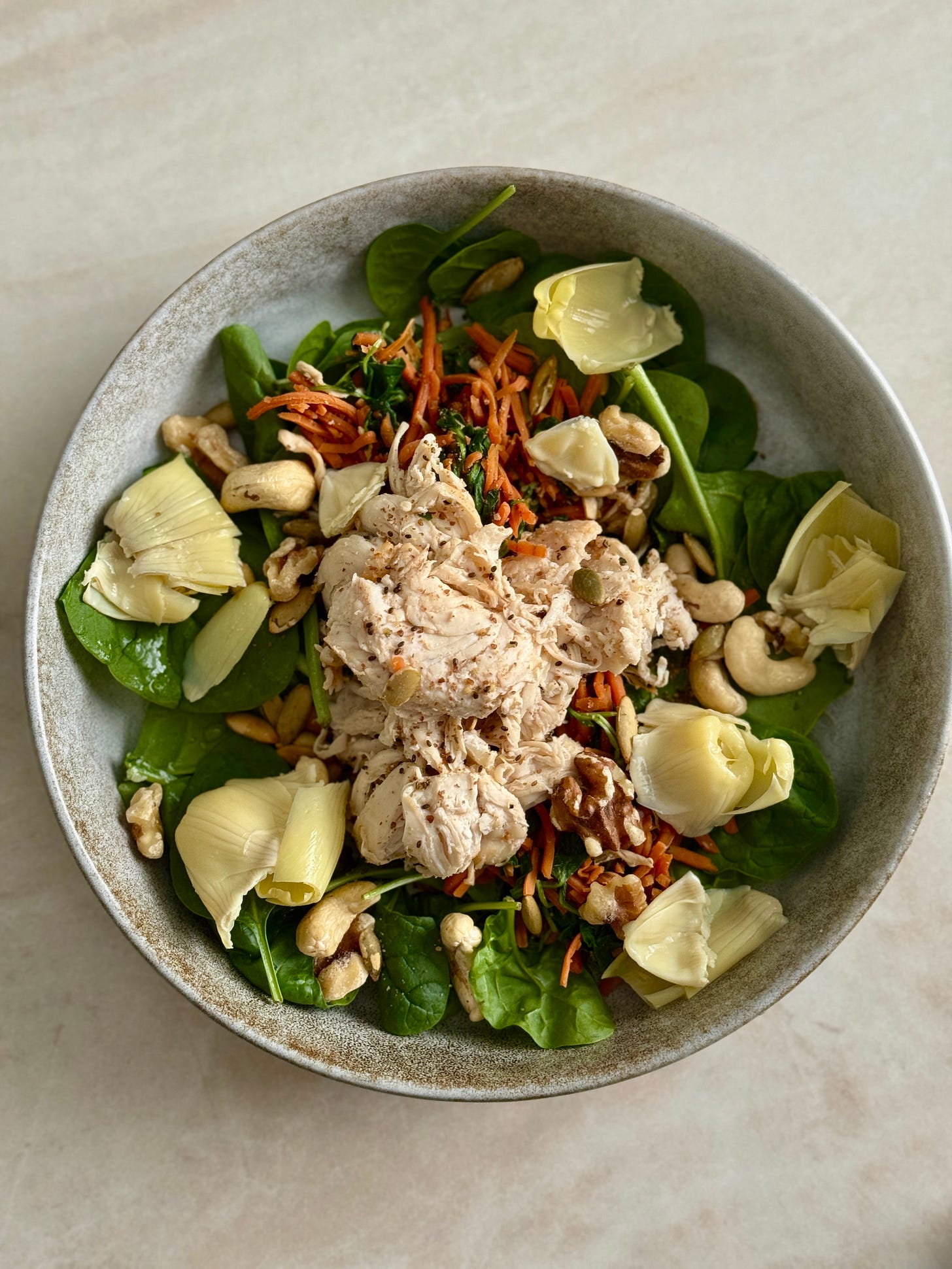 Warm chicken and shredded carrot salad with tender artichoke hearts, baby spinach, and toasted seeds, served as a hearty, nutrient-rich bowl.