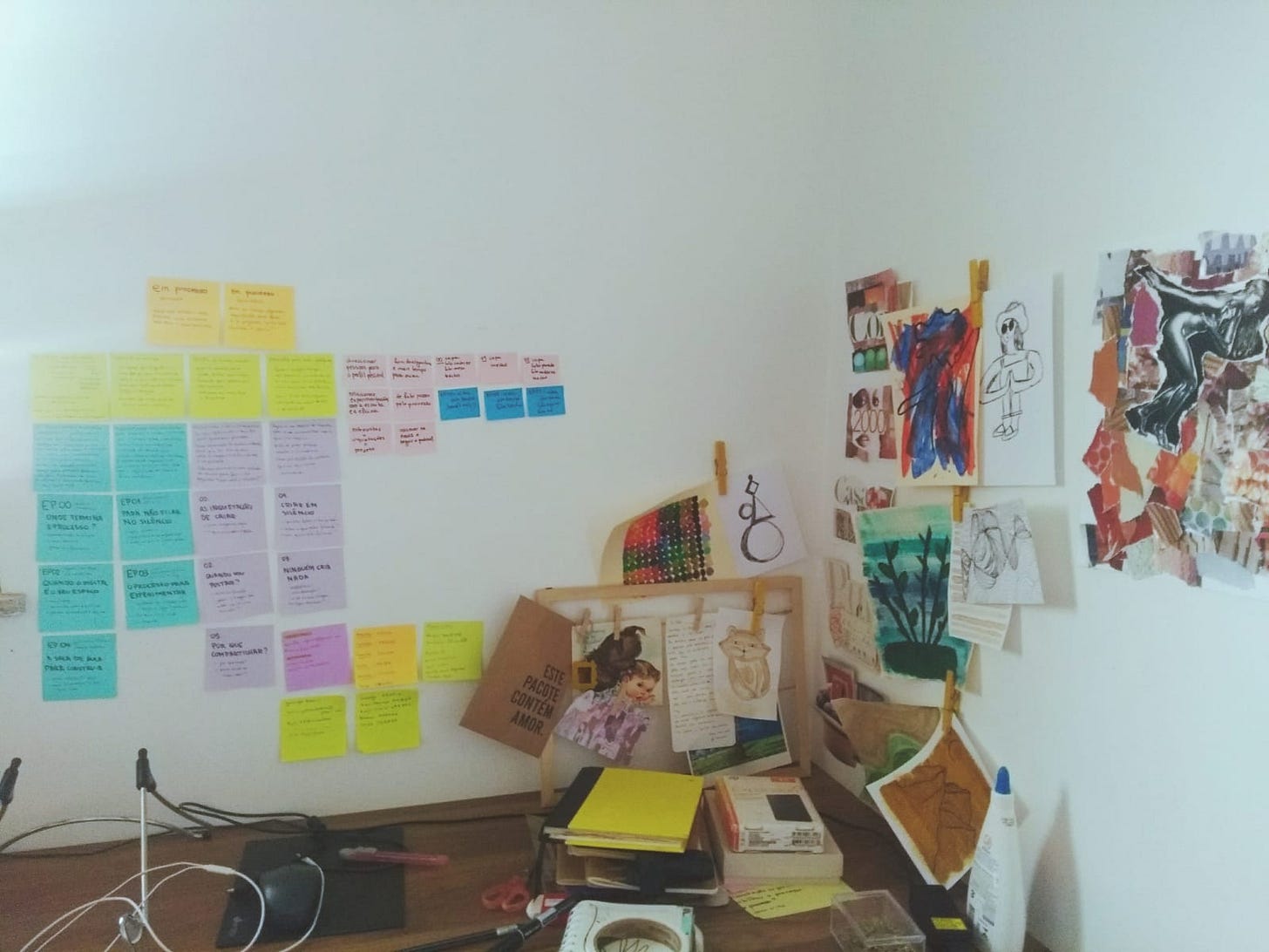 Imagem de uma parede branca com desenhos colados na parede do lado direito. Do ado esquerdo há post-its coloridos. Abaixo há uma mesa de madeira, onde tem cadernos, canetas e papéis.