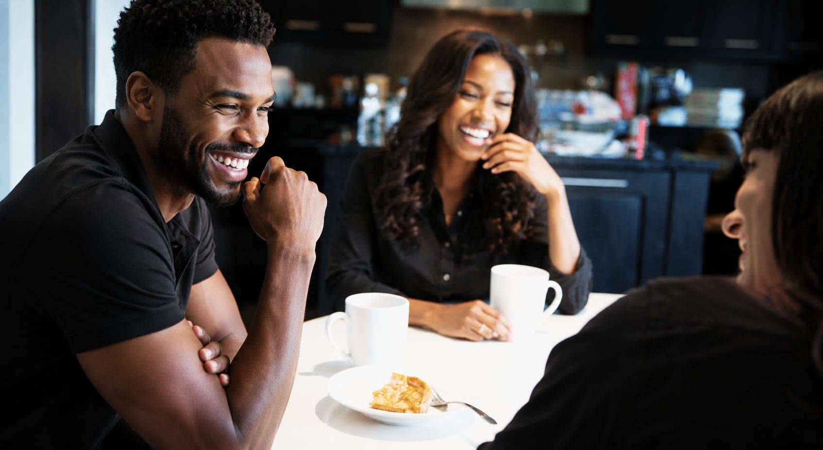 A warm, candid kitchen scene showing two Black couples laughing together around a table with coffee mugs and dessert. The group mirrors the relaxed poses and joyful expressions of a casual lifestyle photo, with soft lighting, dark modern cabinetry, and a cozy, friendly atmosphere. A warm, candid kitchen scene showing two Black couples laughing together around a table with coffee mugs and dessert. The group mirrors the relaxed poses and joyful expressions of a casual lifestyle photo, with soft lighting, dark modern cabinetry, and a cozy, friendly atmosphere.