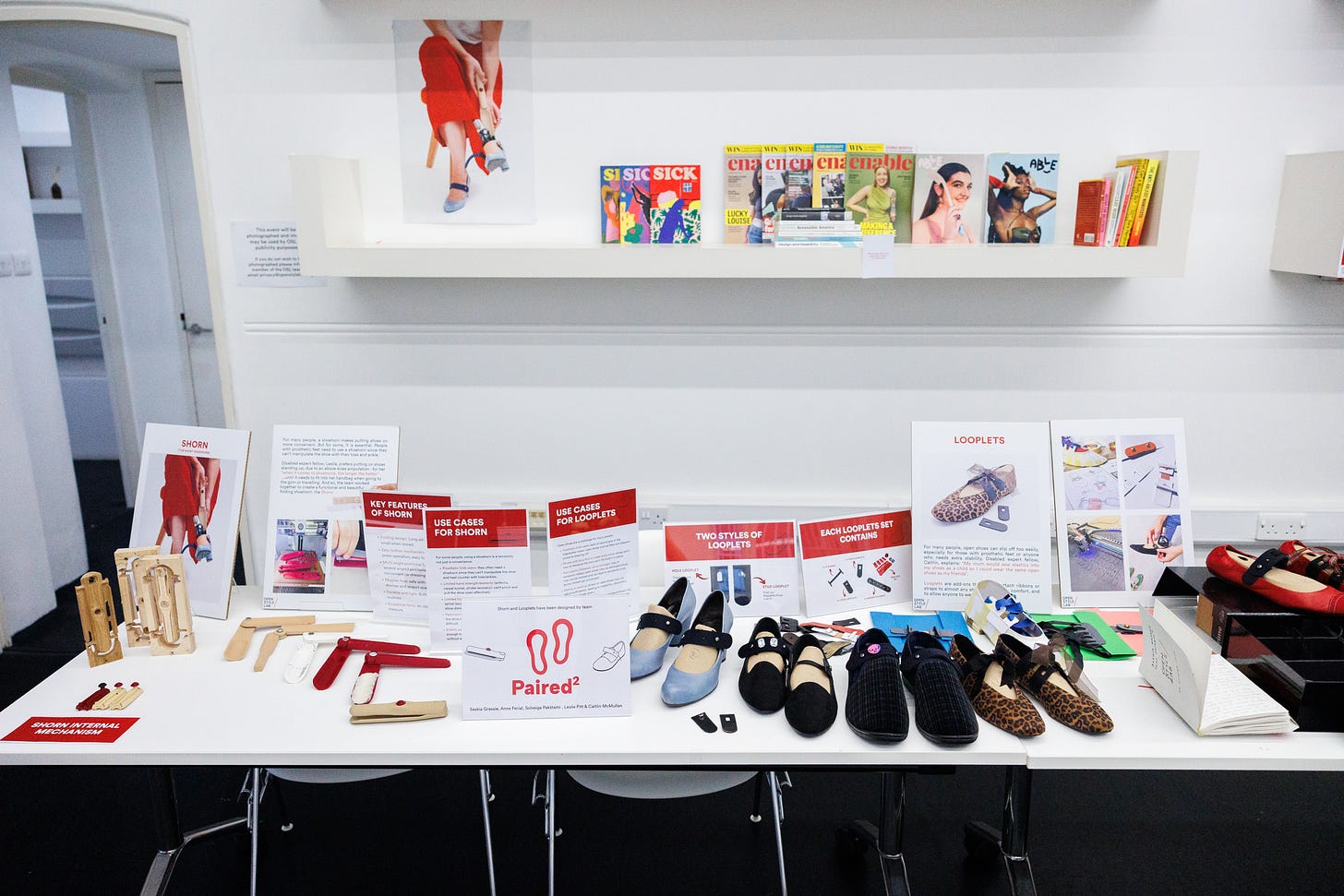 An image of a presentation table. On the table are information cards, alongside ballet-style shoes and heels with attachable straps, and wooden foldable shoe horns. In the background you can see an image posted of someone wearing the shoe design, and a collection of disability media publications