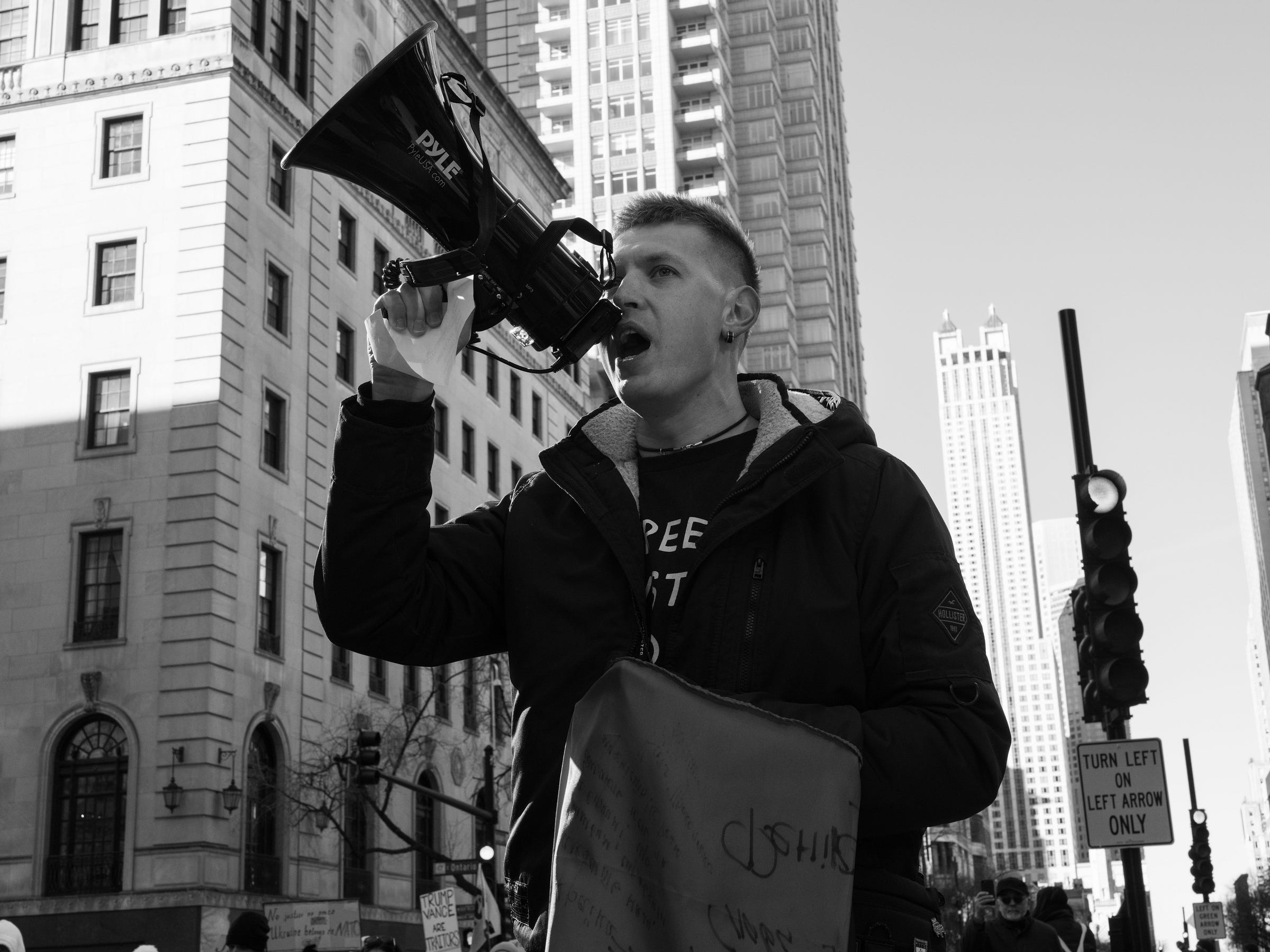 Alt text: A protester passionately speaks into a megaphone during a demonstration in an urban setting. He wears a dark jacket and holds a flag or banner, with tall buildings and traffic lights in the background. His determined expression conveys urgency and activism. Alt text: A protester passionately speaks into a megaphone during a demonstration in an urban setting. He wears a dark jacket and holds a flag or banner, with tall buildings and traffic lights in the background. His determined expression conveys urgency and activism.