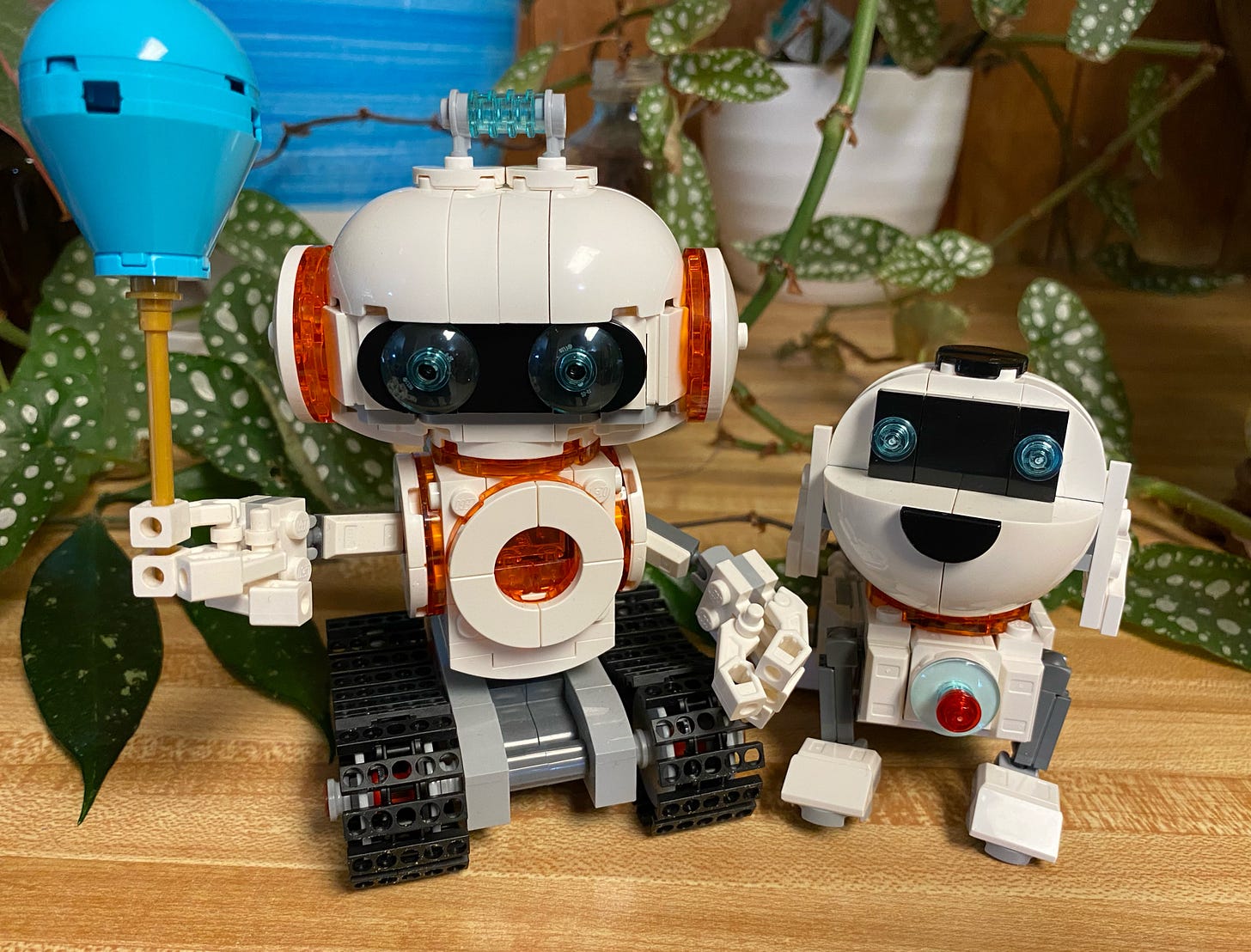 A photograph of two Lego figurines. One is a white and orange robot on treads holding a blue balloon. The other is a white and orange robot dog. They are standing amidst house plants.