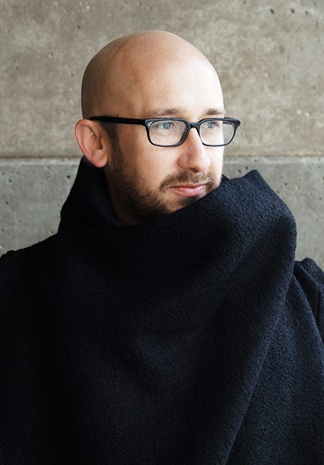 Timothy McCormack standing against a grey stone wall. He is wearing a black woolen coat with a collar that seems to double as a scarf..