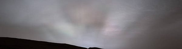 A panoramic image captured by the Curiosity Mars rover. The dark horizon appears as a jagged sliver along the bottom of the image with the gray Martian sky above. Taken at twilight, the image displays faint white, green, and pink-hued “sun rays” shining through clouds at sunset. The varying light and dark beams radiate from the center of the image upward and outward from the horizon. This panorama was stitched together from 28 images sent to Earth. Credit: NASA/JPL-Caltech/MSSS/SSI
