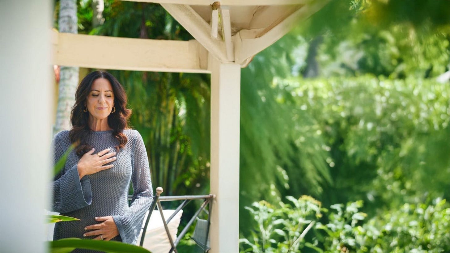 A woman with long dark wavy hair stands on a porch with white wooden beams, wearing a gray knit sweater with bell sleeves. She has one hand resting on her chest and the other on her midsection, eyes closed, in a peaceful pose. Lush green foliage surrounds the porch in the background. A woman with long dark wavy hair stands on a porch with white wooden beams, wearing a gray knit sweater with bell sleeves. She has one hand resting on her chest and the other on her midsection, eyes closed, in a peaceful pose. Lush green foliage surrounds the porch in the background.