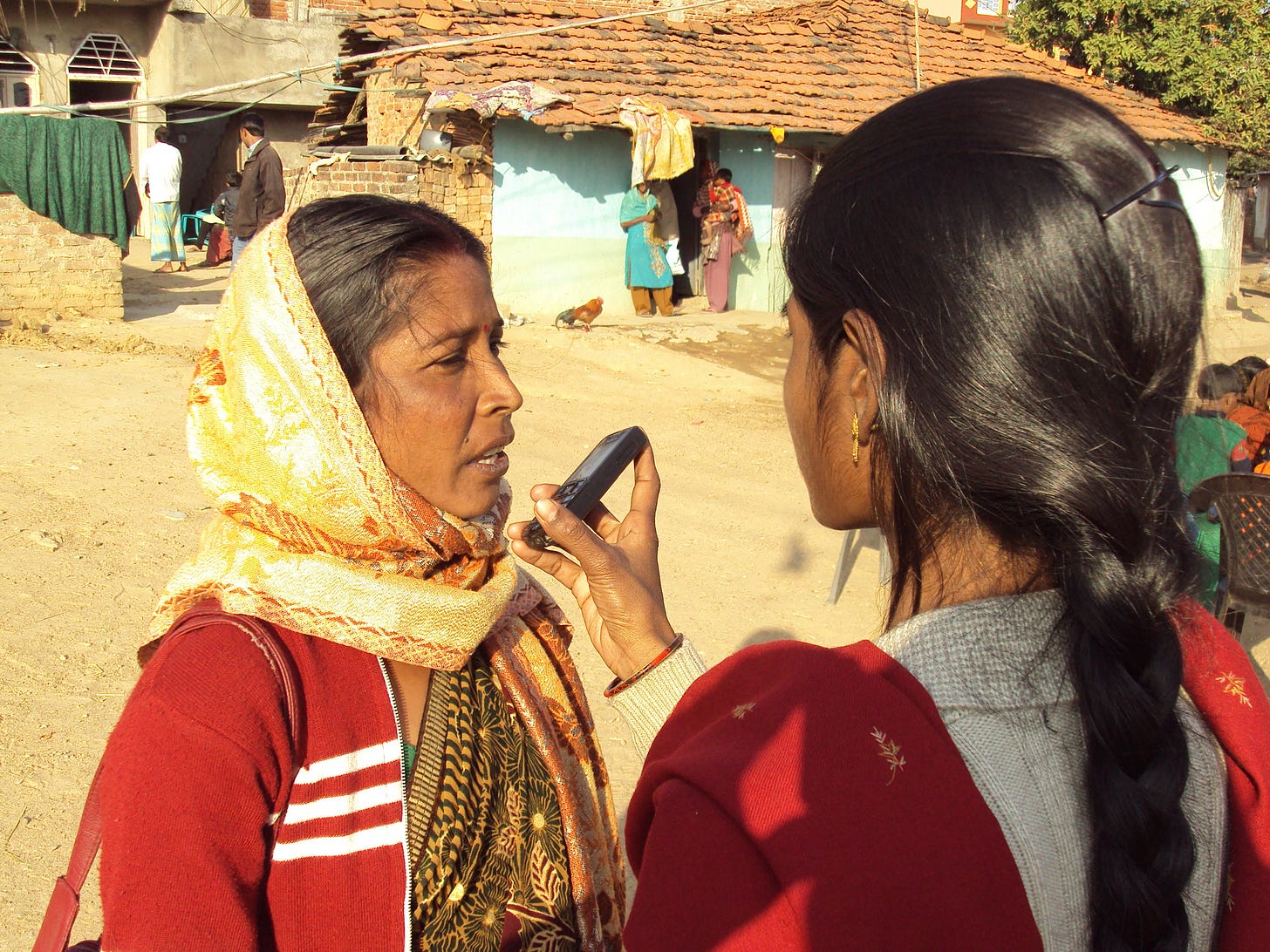 A woman holding a feature phone next to a middle aged lady in a village. The woman holding the phone is demonstrating Mobile Vaani to the lady