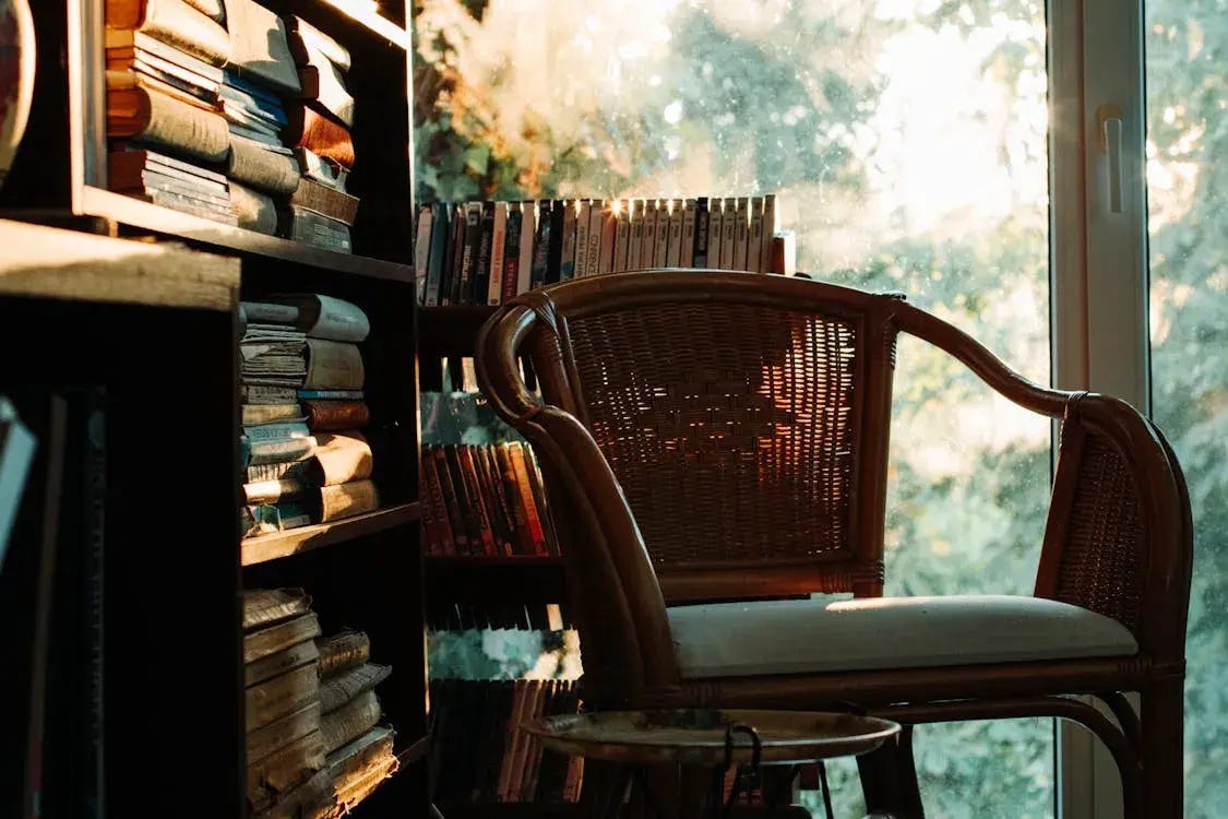 Wicker reading chair beside tall bookshelves and a sunlit window, creating a warm and cozy library corner.