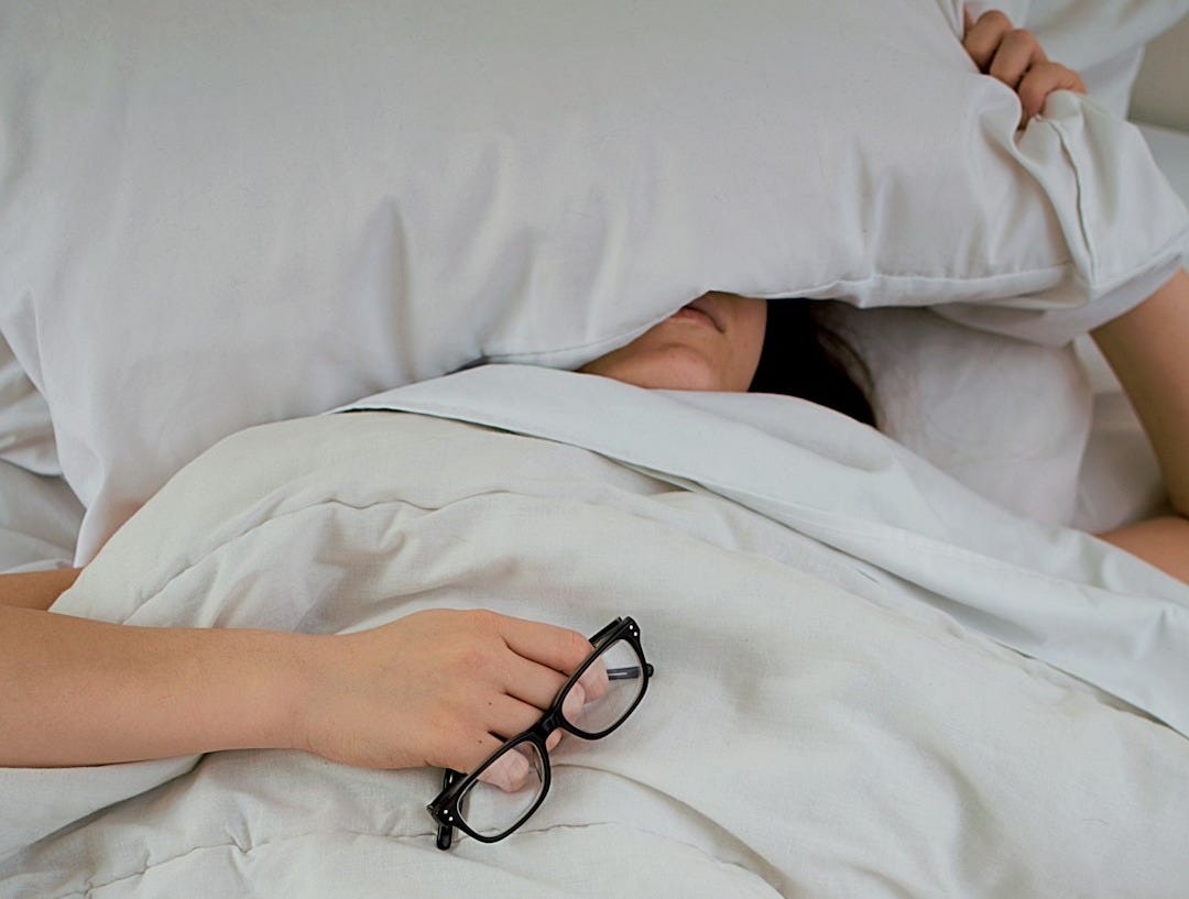 person lying on bed while covering face with pillow and holding eyeglasses