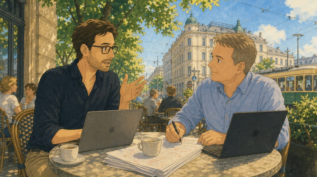 Two men sit at a café table in Helsinki on a sunny day, coffee cups, laptops and printed pages in front of them. The man on the left leans forward gesturing as he speaks. The man on the right holds a pen and listens. Behind them, classical Helsinki architecture and a tram are visible beneath summer foliage.