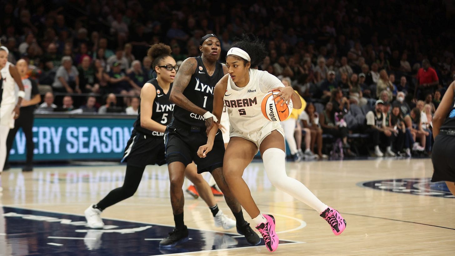 A basketball player in a white Dream jersey numbered 5 dribbles an orange ball past two defenders in black Lynx jerseys on an indoor court, with a crowd in the stands behind them. A basketball player in a white Dream jersey numbered 5 dribbles an orange ball past two defenders in black Lynx jerseys on an indoor court, with a crowd in the stands behind them.