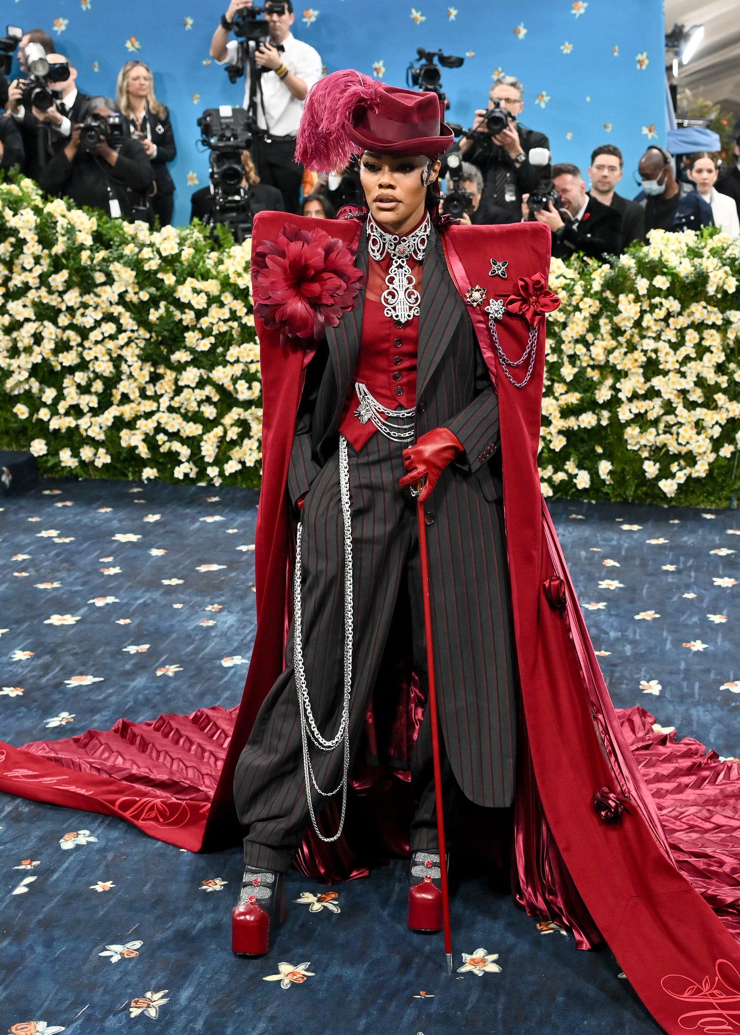 Teyana Taylor in an elaborate pinstriped suit and red cloak with dramatic shoulders, a cane and a feathered hat