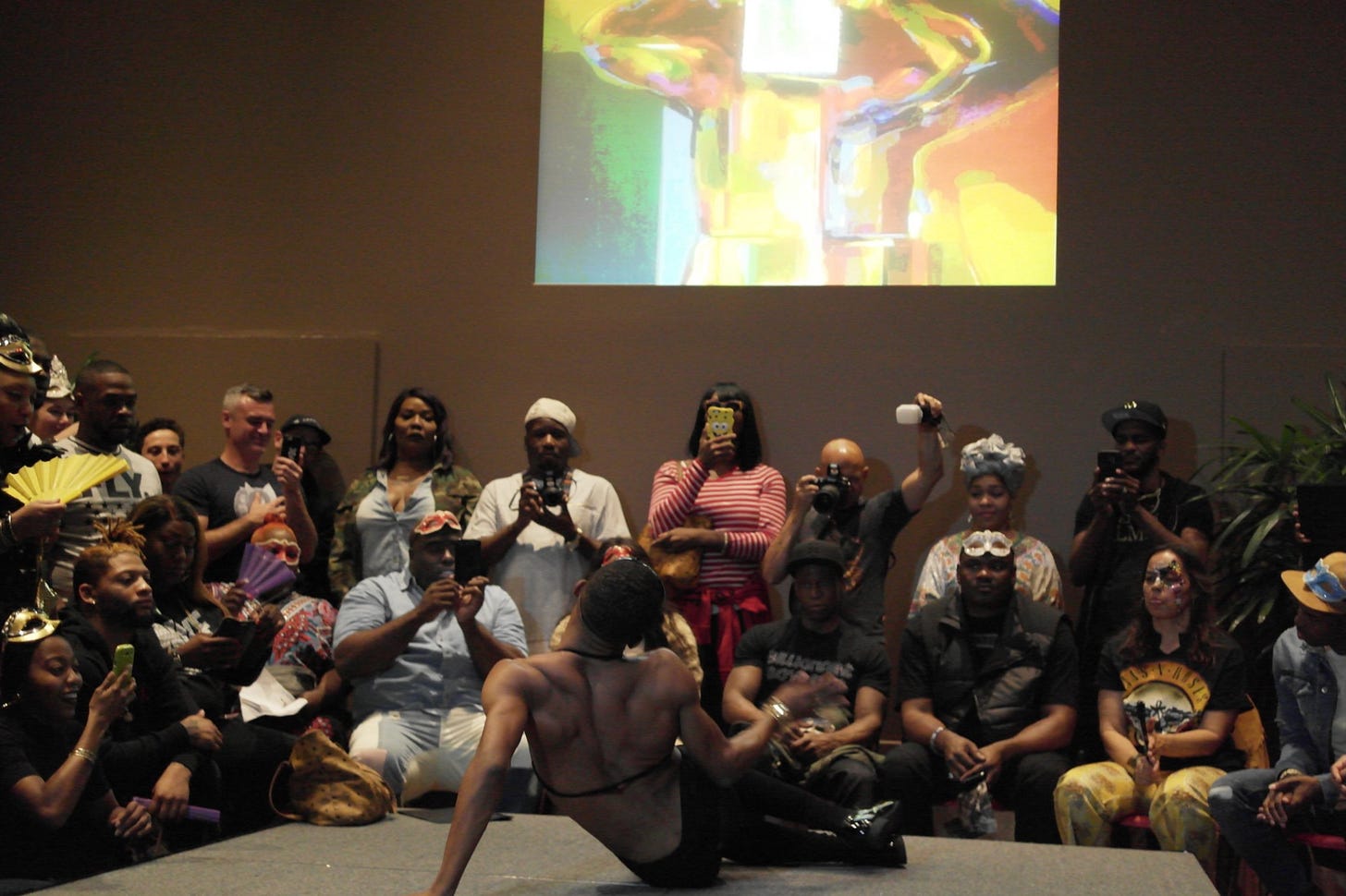Photo taken from behind a ballroom performer seated at the end of a stage in a dance pose. An audience of primarily Black people with some dressed in ballroom attire watches attentively. Photo taken from behind a ballroom performer seated at the end of a stage in a dance pose. An audience of primarily Black people with some dressed in ballroom attire watches attentively.