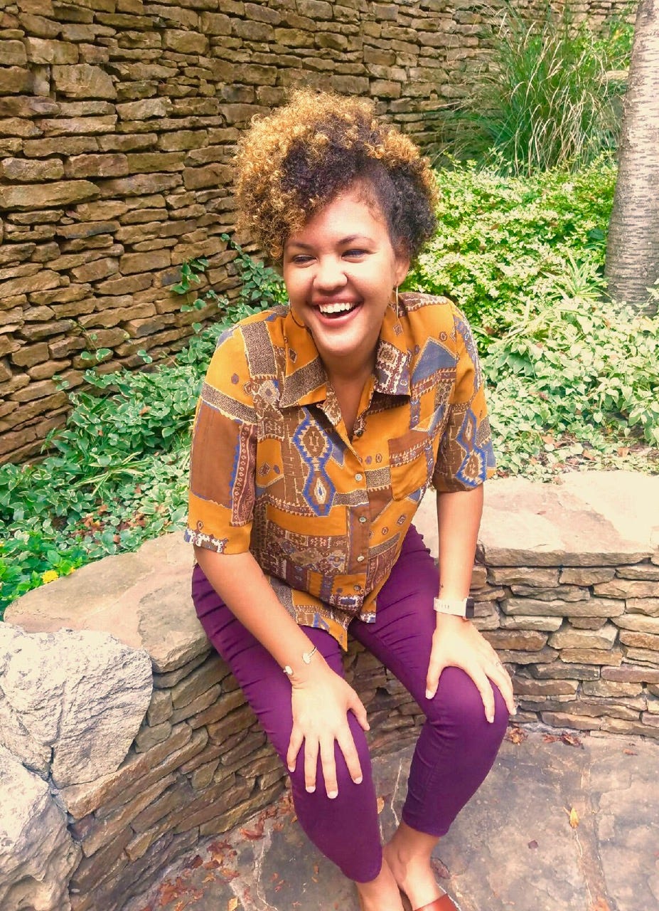 A black woman smiles, sitting on a stone bench with green shrubbery behind her. She is wearing an orange shirt with a graphic design and purple pants. 