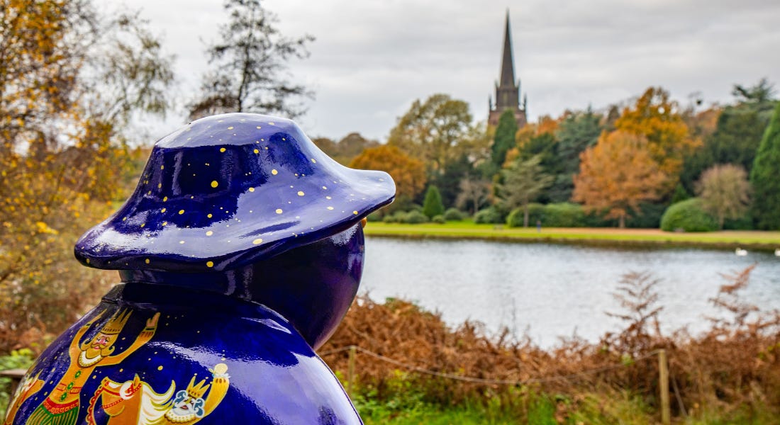 A dark blue Snowman statue facing a river with a church spire in the distance
