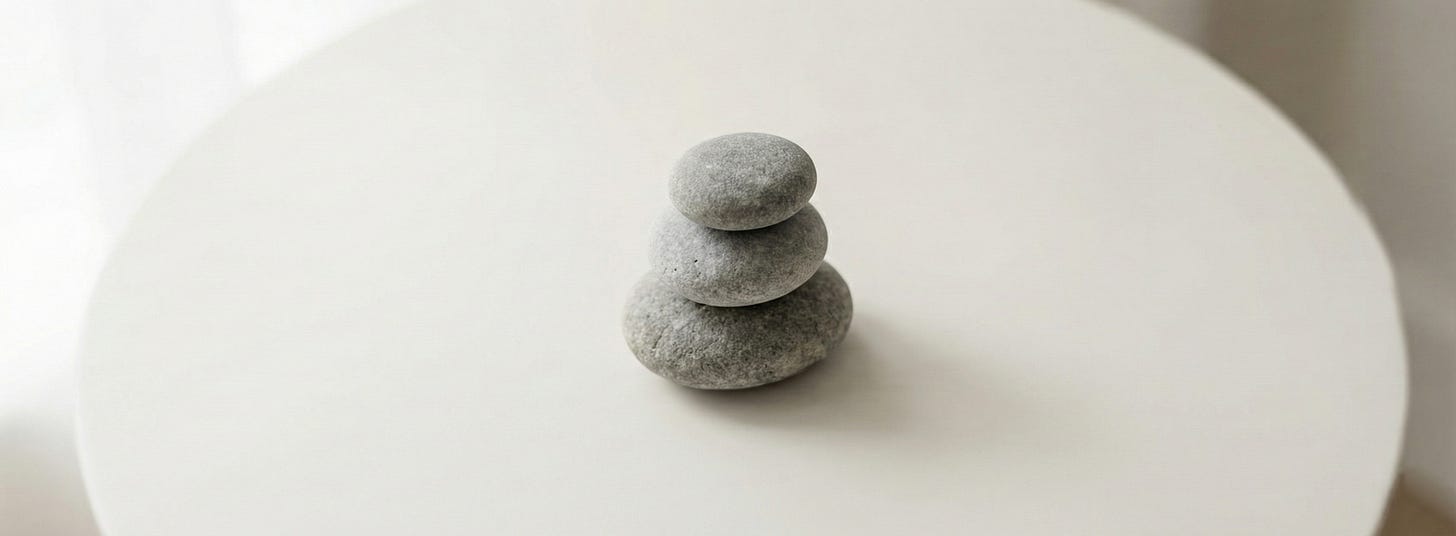 Three smooth grey river stones stacked in balanced cairn formation on round white pedestal table with soft diffused natural lighting. Three smooth grey river stones stacked in balanced cairn formation on round white pedestal table with soft diffused natural lighting.