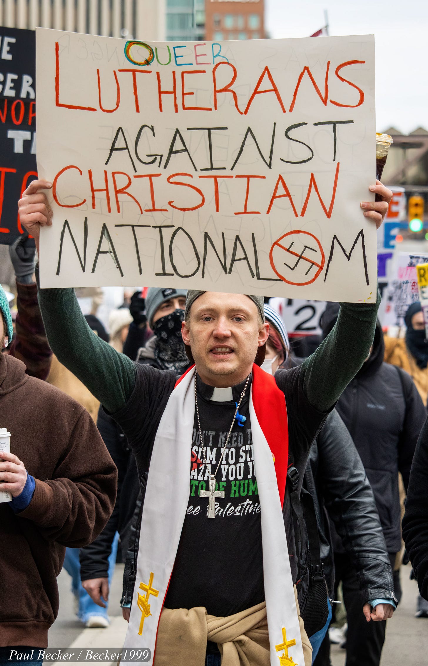 A person at a street protest holds a large handmade sign reading “Queer Lutherans Against Christian Nationalism,” with a rainbow-colored letter in “Queer” and a no symbol over part of the word “Nationalism.” The person wears a clerical-style collar, a long white stole with gold crosses, and layered clothing, while a crowd of bundled-up protesters marches behind them in an urban setting.