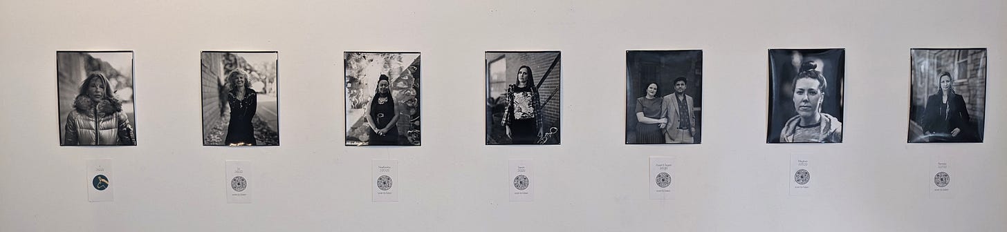 Seven black and white portraits hang on a while wall with labels underneath Seven black and white portraits hang on a while wall with labels underneath