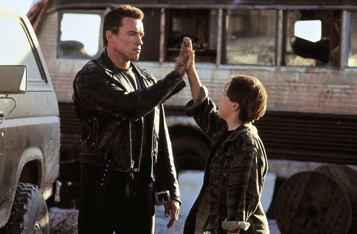 a frame from the movie Terminator 2 showing Arnold Schwarzenegger high fiving the teenaged boy John Connor in front of a dilapidated bus