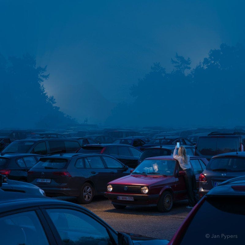 A woman stands outside a red car in a dimly lit parking lot at dusk, using her phone. The car’s headlights are on, and the lot is filled with other parked vehicles under a misty blue sky. A woman stands outside a red car in a dimly lit parking lot at dusk, using her phone. The car’s headlights are on, and the lot is filled with other parked vehicles under a misty blue sky.