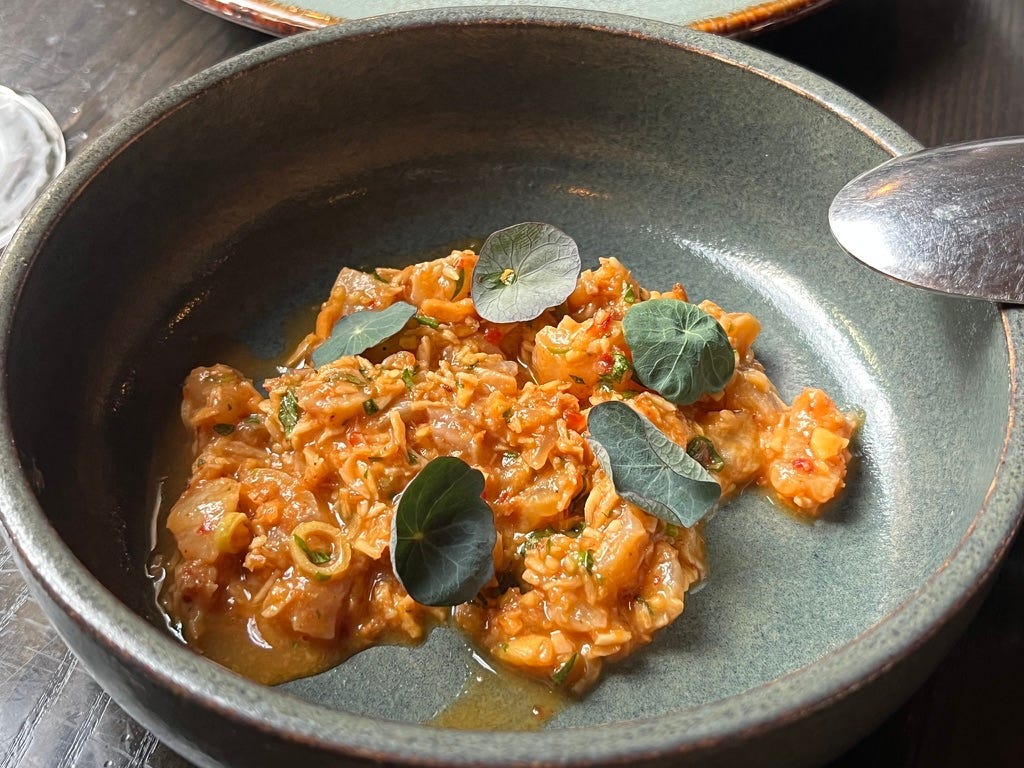 pieces of white fish with other small pieces of food, covered in orange sauce, with a few green leaves on top, all on a round green plate