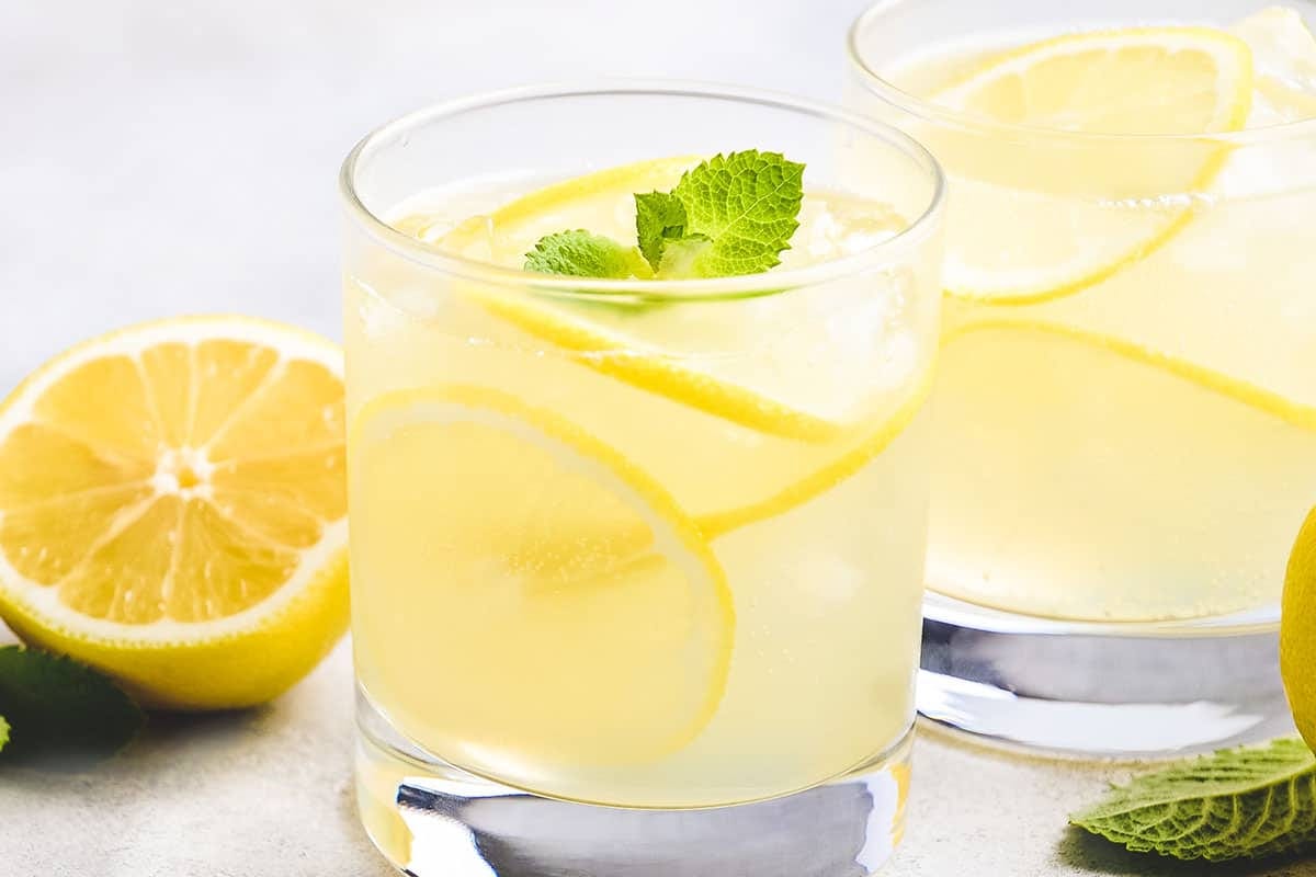 Homemade hard lemonade served over ice with fresh lemon slices and mint, with a pitcher of lemonade in the background.
