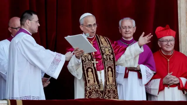 Pope Leo speaks into a microphone surrounded by other men in religious clothing. He speaks from the balcony of St. Peter's Basilica with his arms widened. Pope Leo speaks into a microphone surrounded by other men in religious clothing. He speaks from the balcony of St. Peter's Basilica with his arms widened.
