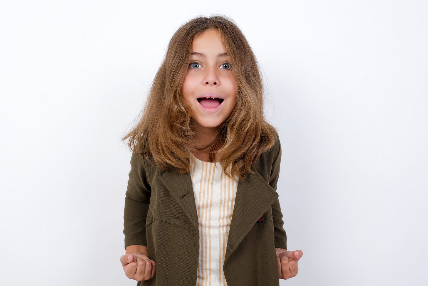 Close up portrait of stylish modern little girl standing against white background with wave hairstyle raising fists up isolated on shine grey background big eyes and wide open mouth
