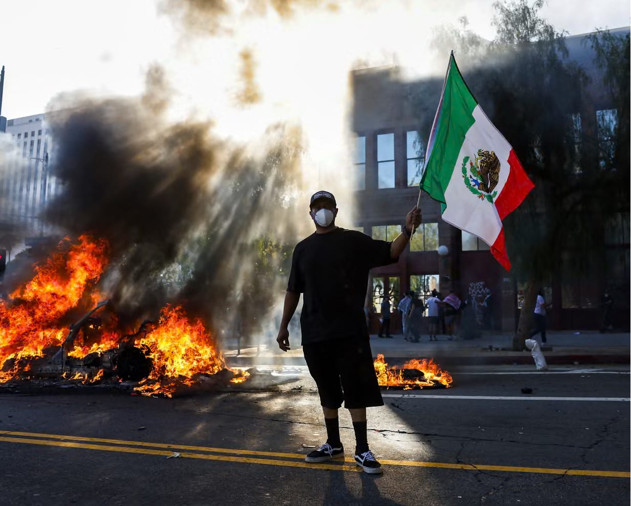 A protester in Los Angeles, holding a Mexican flag with a burning car in the background. A protester in Los Angeles, holding a Mexican flag with a burning car in the background.