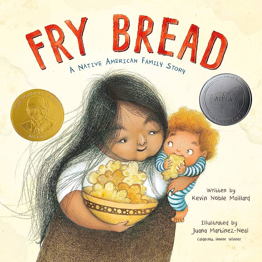 The cover of  Fry Bread: A Native American Family Story written by Kevin Noble Maillard and illustrated by Juana Martinez-Neal. The cover depics an older native woman with long, flowing black hair holding a bowl of fry bread in one arm and a baby in blue pajamas with yellow curly hair who is eating a piece of fry bread in her other arm.                                                                                                                                La portada de Fry Bread: A Native American Family Story escrita por Kevin Noble Maillard e ilustrada por Juana Martinez-Neal. La portada muestra a una mujer nativa mayor con cabello negro largo y suelto que sostiene un plato de pan frito en un brazo y un bebé en pijama azul con cabello amarillo rizado que está comiendo un trozo de pan frito en el otro brazo.