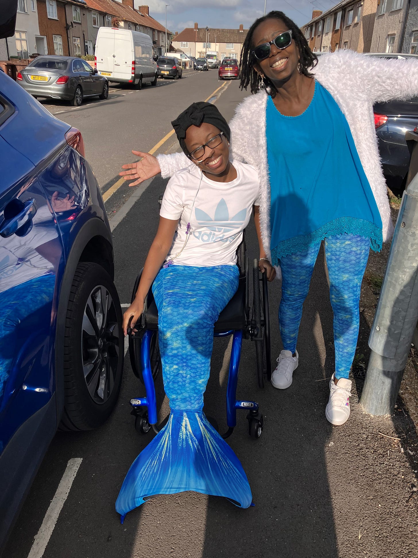 Very similar to the previous picture, except the sun is on them both. Cyrene is squinting due to this. She is also leaning on her wheelchair with both hands, one on the brake handle, and one on the push rims of her wheels.