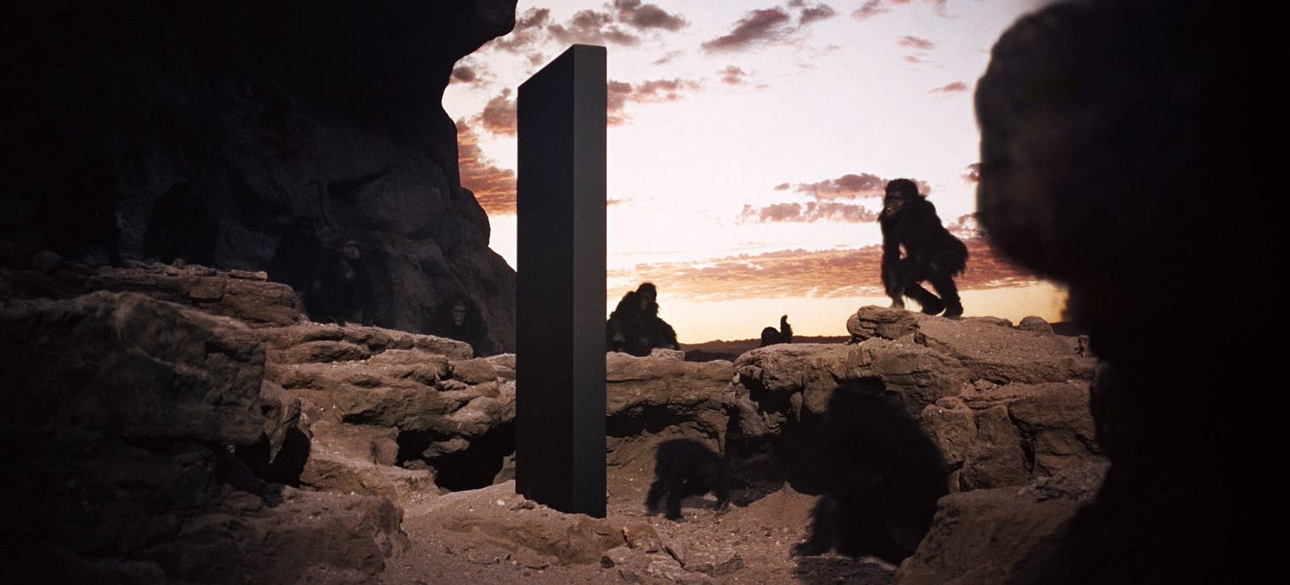 A group of apes surround a strange monolith in the middle of a rock landscape.
