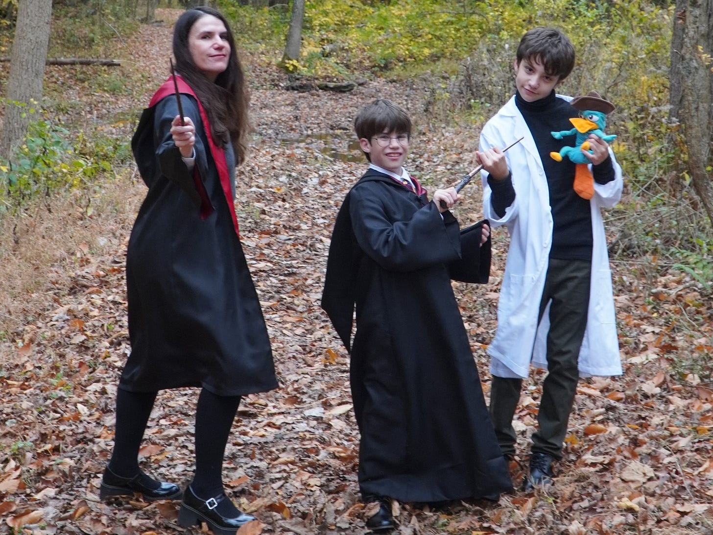 A woman dressed like Hermione Granger stands next to a boy dressed like Harry Potter and a boy dressed like Dr. Doofenshmirtz. They are in the woods. It’s fall and daytime. 