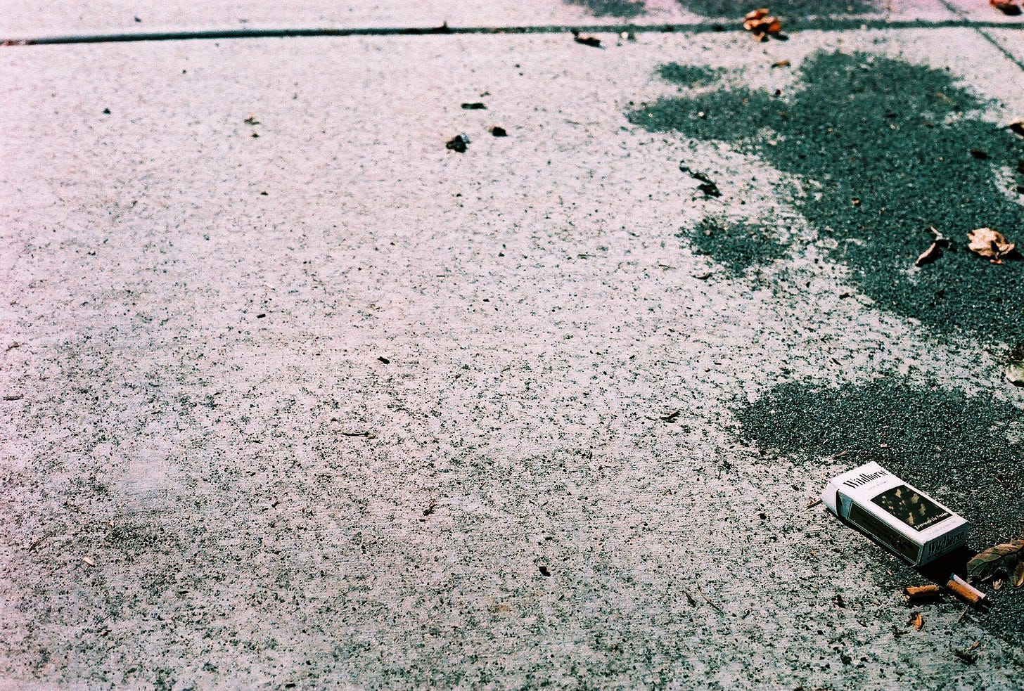 A film photograph of the sidewalk. On the righthand side of the frame lies a box of cigarettes and some gravel / asphalt that looks like tree shadows.