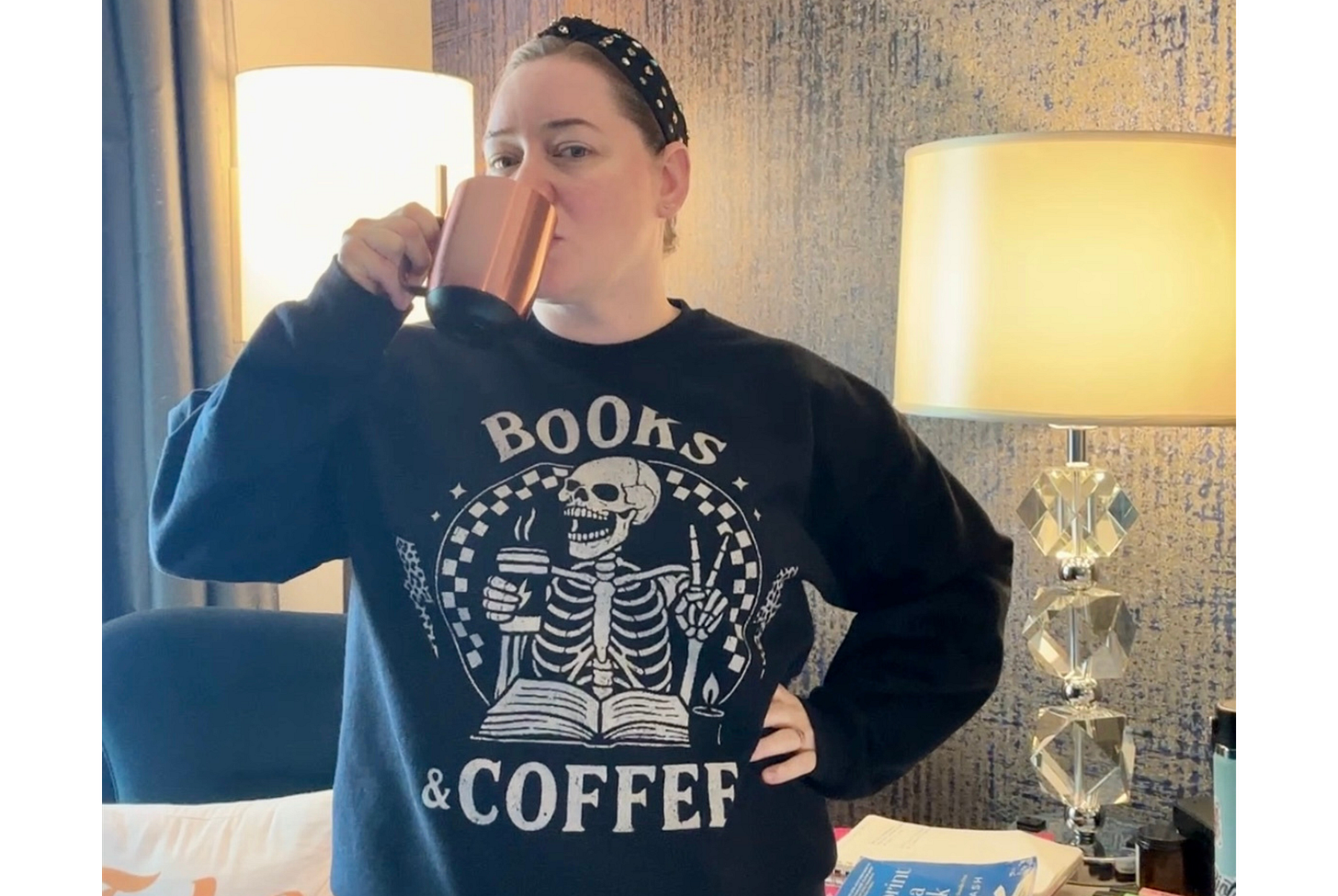 Candice sips coffee from a rose gold coffee mug. Hair held back with a rhinestone studded black headband. Wearing a black sweatshirt with a book reading skeleton who is holding a coffee cup and throwing up a peace sign. Candice sips coffee from a rose gold coffee mug. Hair held back with a rhinestone studded black headband. Wearing a black sweatshirt with a book reading skeleton who is holding a coffee cup and throwing up a peace sign.
