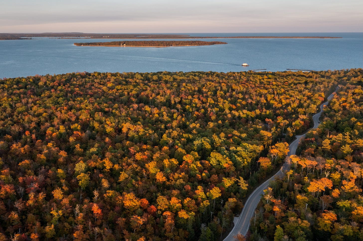 Aerial view of a winding forest road lined with red, orange, and yellow autumn trees in Door County, Wisconsin, with Lake Michigan in the distance.