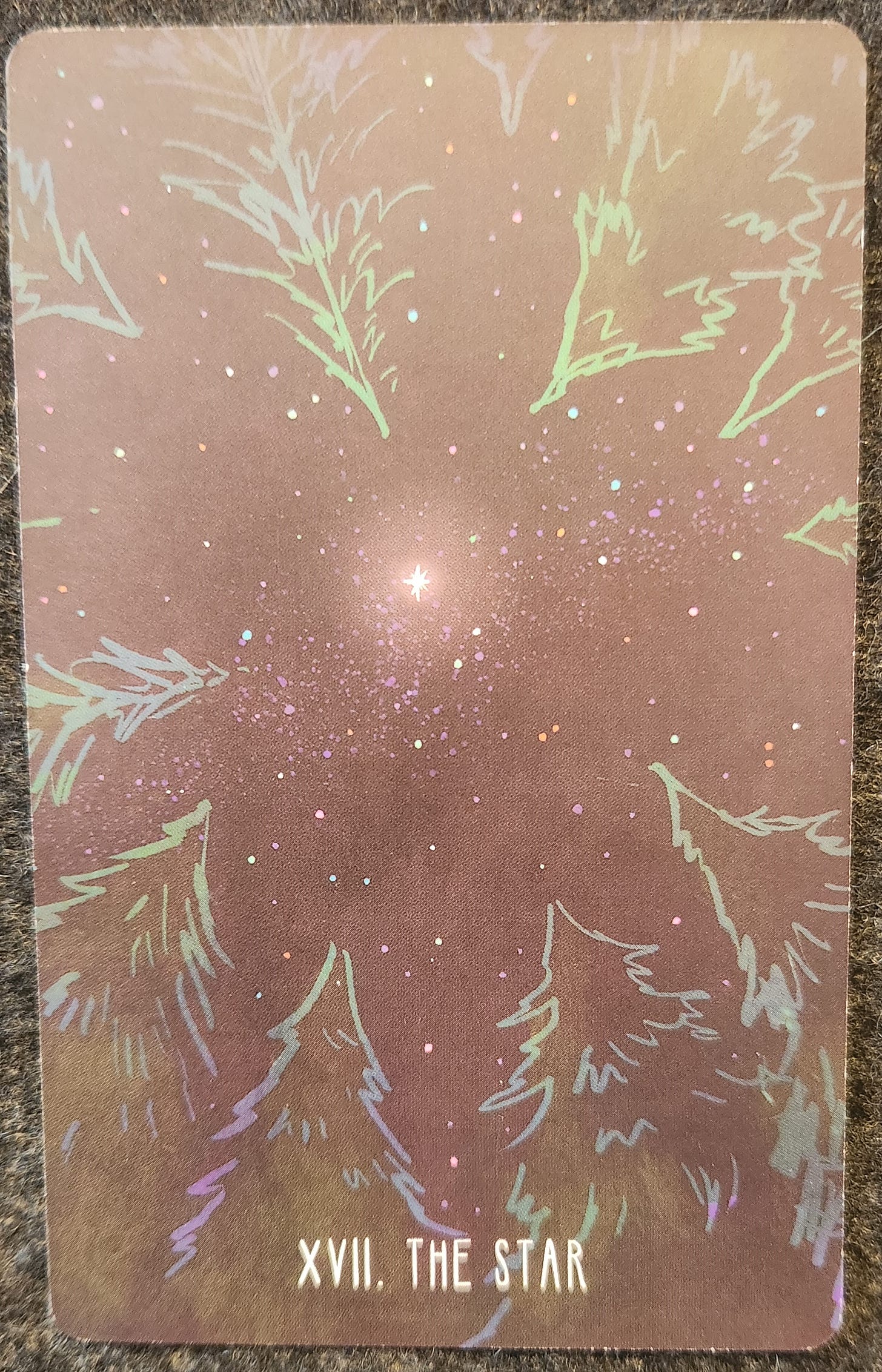 A tarot card labelled XVII. The Star. A bright star shines in the middle of a starry night sky framed by a clearing of pine trees. The tips of the trees are all pointed towards the star. 
