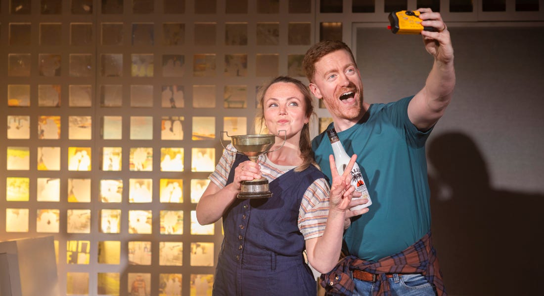 A woman in a blue jumpsuit and stripy t-shirt and a man in a green t-shirt stand in front of a wall. The woman holds a silver trophy cup and has her left hand fingers in a peace sign. The man holds a bottle of Malibu in his right hand and a disposable camera in his left. He is taking a selfie photo of them both, as they smile towards the camera. In the background, a wall is covered in a white grid pattern. A woman in a blue jumpsuit and stripy t-shirt and a man in a green t-shirt stand in front of a wall. The woman holds a silver trophy cup and has her left hand fingers in a peace sign. The man holds a bottle of Malibu in his right hand and a disposable camera in his left. He is taking a selfie photo of them both, as they smile towards the camera. In the background, a wall is covered in a white grid pattern.