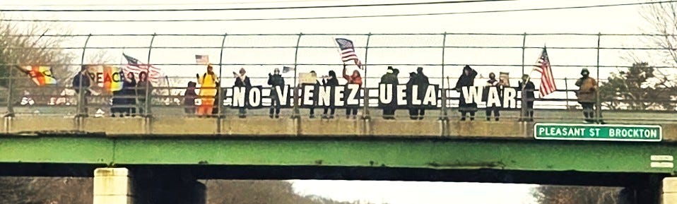 Group of people on an overpass spelling “NO VENEZUELA WAR!” while waving U.S. flags; street sign reads Pleasant St, Brockton.
