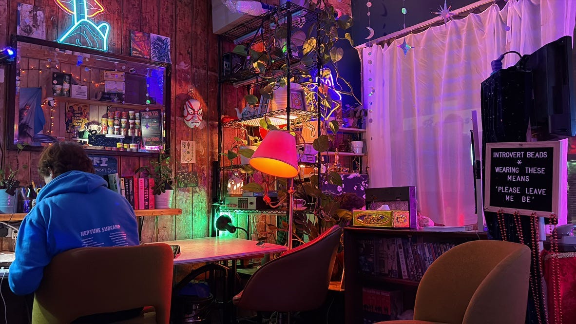A person in a blue sweatshirt sits at a desk, facing away from the camera. It's night time and the room is aglow with neon lights, lamps, twinkle lights. A person in a blue sweatshirt sits at a desk, facing away from the camera. It's night time and the room is aglow with neon lights, lamps, twinkle lights.