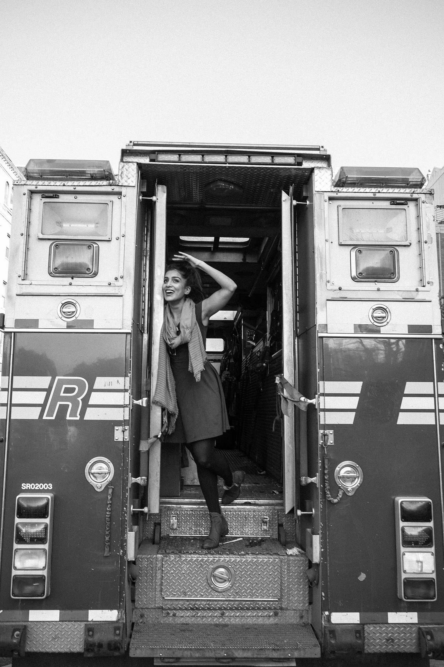 A black and white photo of actress Pallavi Sharda posing playfully in the open doorway at the back of a fire truck. She is smiling with one hand in her hair, framed by the vehicle's large metal doors.