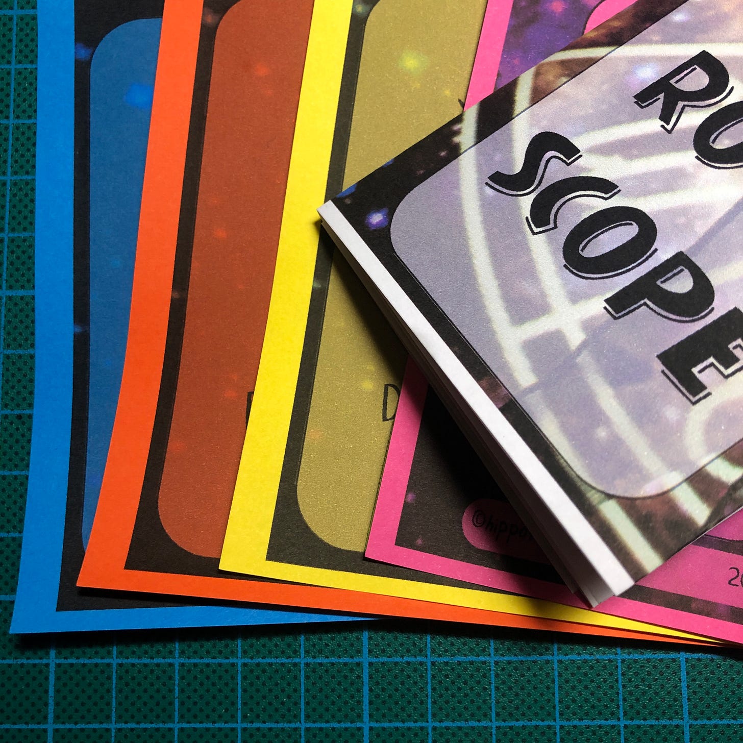 Photo du nouveau zine imprimé sur des feuilles de différentes couleurs. Celui en papier blanc est plié, les autres sont à plat.