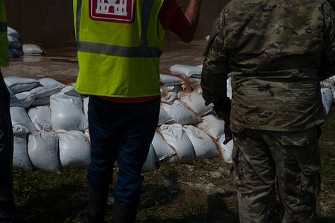 The progression of a sand boil from occurrence to remediation: 1) A soldier in fatigues carries a sandbag downhill alongside a floodway structure (a tall wall) with a cigarette between their lips and eyes turned down toward the target in the grass. The sand boil looks like a gurgling fountain a few inches in diameter, with muddy water spreading around it. 2) The floodway structure, chain of soldiers and ring dyke (several dozen sandbags stacked around the sand boil in a half-moon formation at the base of the wall) are seen from just uphill. A person in a neon yellow safety vest stands in muddy, ankle-deep water as it rises inside the ring dyke, which is now a few sandbags high. Inside the floodway structure, floodwaters as sludgy as brownie mix appear to loom several feet over the heads of the soldiers working on the landside. Two military SUVs pass on the road in the background. 3) Soldiers in fatigues pass sandbags down from pallets on a flatbed. In the foreground, a blond soldier with a square jaw and solemn expression looks on in a camouflage cap. 4) Soldiers in fatigues stand perpendicular to the floodway structure, passing sandbags toward the ring dyke. 5) Two people stand side by side, facing the ring dyke with their backs to the photographer. Muddy water spills over the sides of the ring dyke, which now stands five or six sandbags high. The person on the right is in fatigues. The person on the left wears a neon yellow safety vest with the red and white turreted castle emblem of the U.S. Army Corps of Engineers. 6) Several members of the Oklahoma National Guard stand in, on and around the ring dyke in fatigues. One is submerged to calf- or knee-height in the muddy water spilling over the edges of the ring dyke. Another steps calf-deep into the pool with one leg, straddling the ring dyke to position a sandbag.