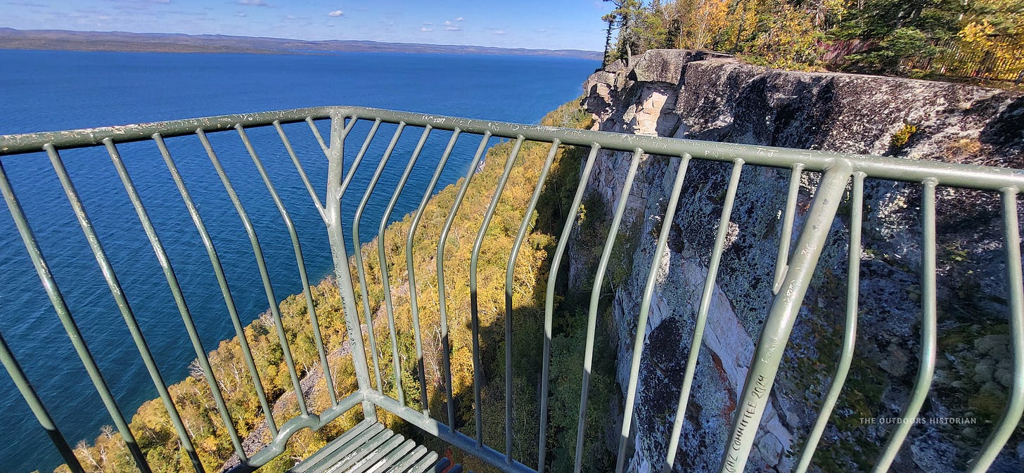 A panoramic view from the Thunder Bay Lookout at Sleeping Giant Provincial Park. To the right, jagged cliffs drop off into a steep talus slope covered in hardy green vegetation. To the left, the vast, deep blue expanse of Lake Superior meets the horizon under a bright sky.