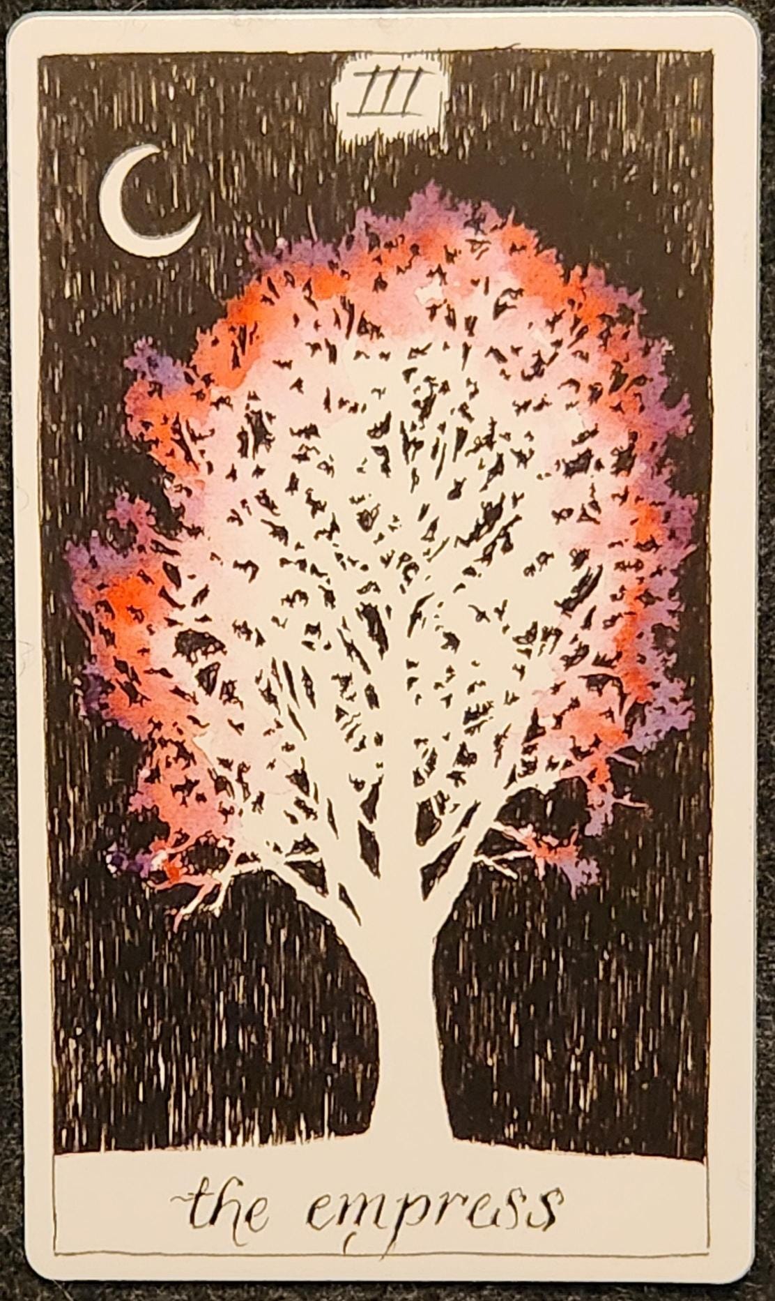 A tarot card labelled III the empress. A white tree with full leaves and branches. The edges of the leaves are pink and purple, which are the only colours in the image. The sky above the tree is black, with a bright crescent moon. 