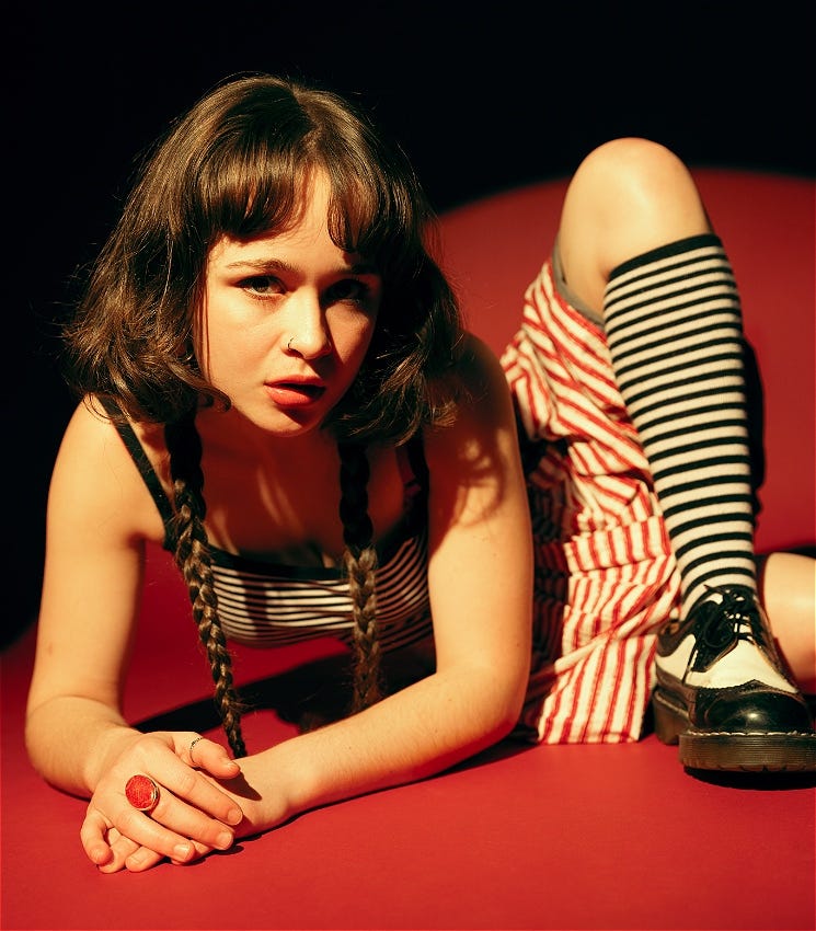 Debby is lying on a bright red background, wearing stripy clothing and looking at the camera. Her body is slightly contorted, with her left leg, wearing a black and white striped sock, as her knee is propped close to her head. She has dark hair with two tapering braids falling on either side of her face.