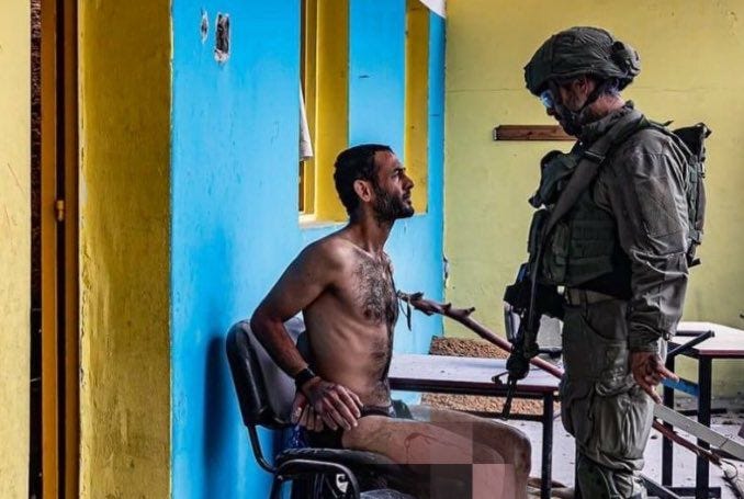 Before the breaking point — An IDF soldier stands over a bound Palestinian civilian, the human body reduced to tactical cover. In the logic of Thanacide, this is not a man with rights or a family; he is a ‘mosquito’ to be used and discarded. Before the breaking point — An IDF soldier stands over a bound Palestinian civilian, the human body reduced to tactical cover. In the logic of Thanacide, this is not a man with rights or a family; he is a ‘mosquito’ to be used and discarded.